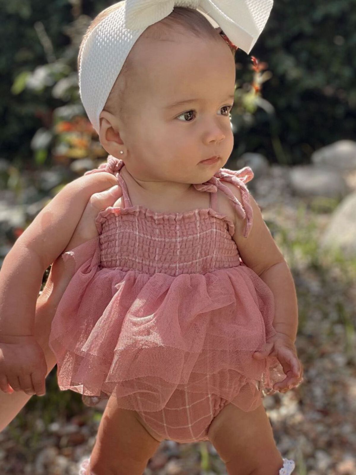 Toddler in a pink outfit with a bow headband, standing outdoors with greenery in the background.