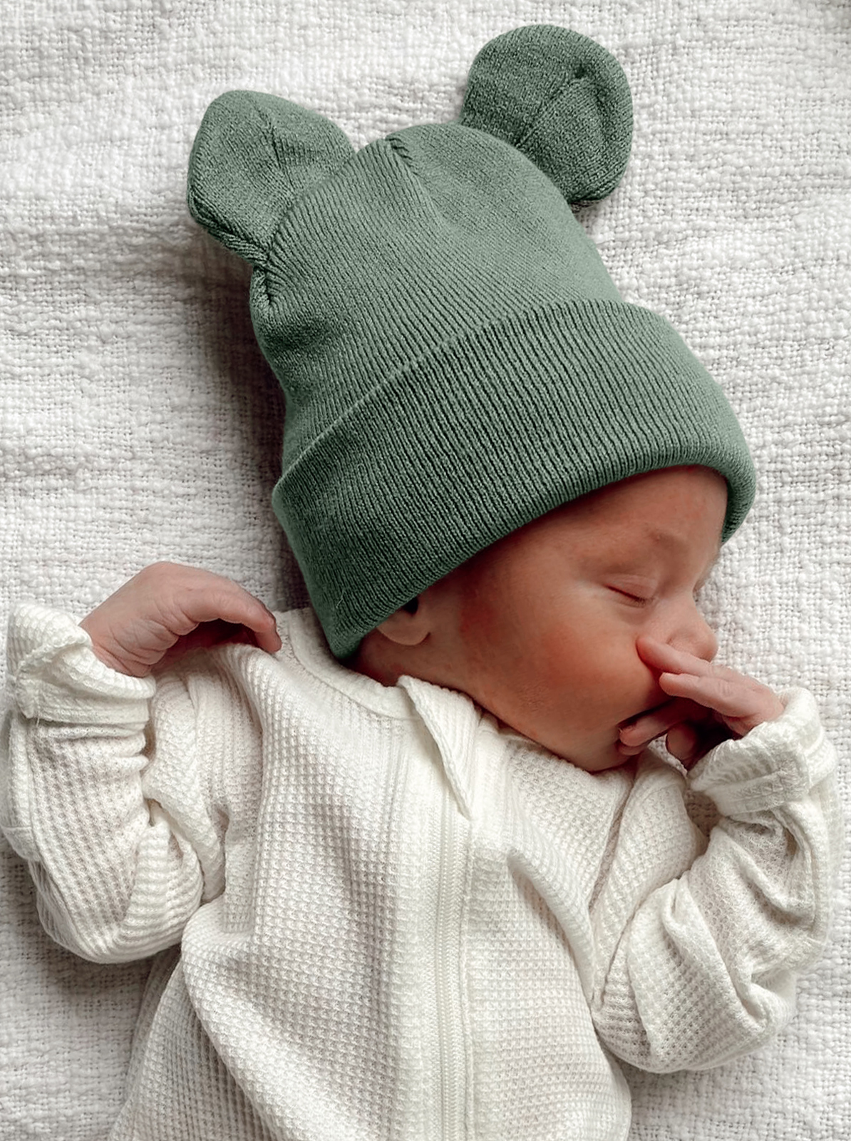 A sleeping baby in a green bear-ear beanie and white outfit, lying on a light textured blanket.