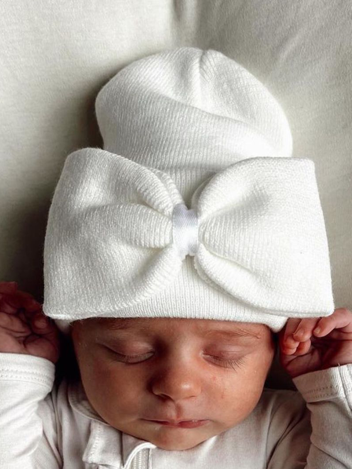 Sleeping baby in a white hat with a large bow, peacefully resting on a light background.