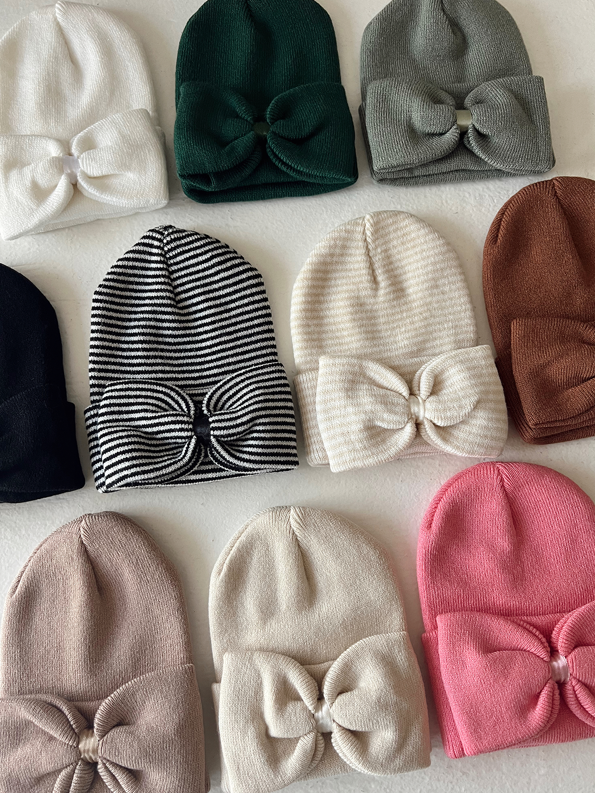 Colorful beanie hats with bows, arranged in a grid on a light background.