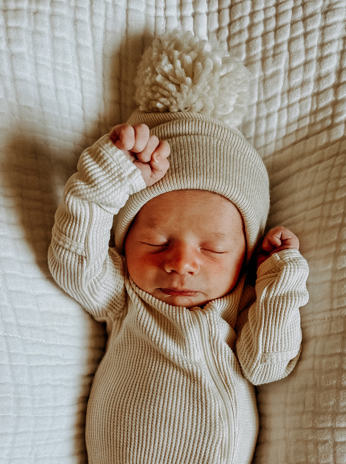 Newborn baby wearing a cream ribbed outfit and a fluffy pom-pom hat, sleeping on a quilted surface.
