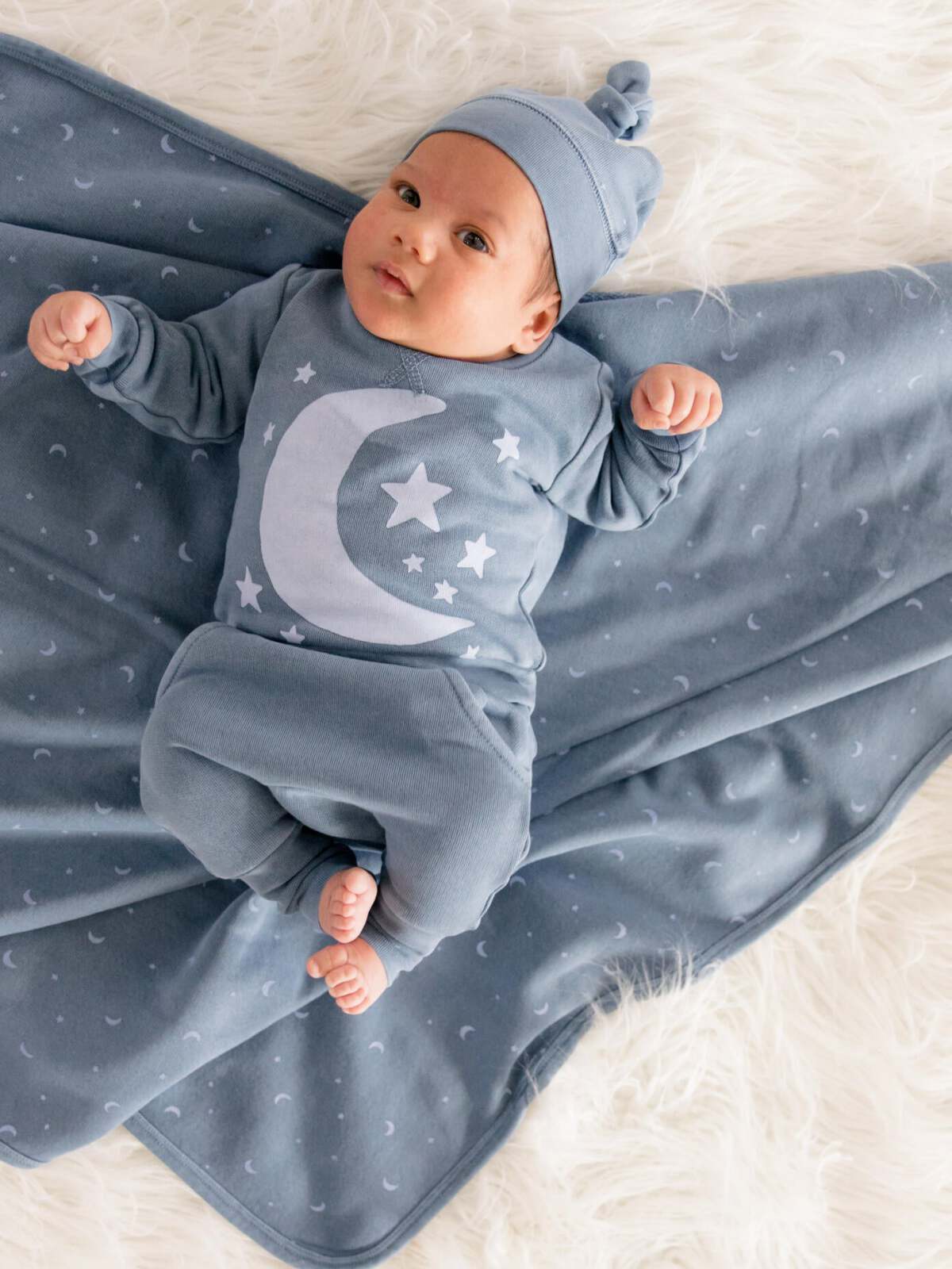 Baby in a blue moon and stars outfit lying on a soft white blanket with a peaceful expression.