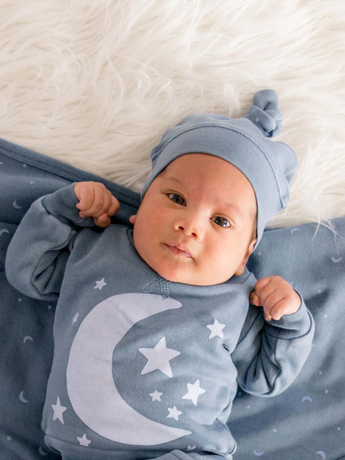 A baby in a blue outfit with stars and a moon lays on a soft, fluffy surface, gazing curiously at the camera.
