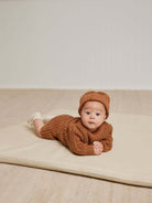 Baby in a cozy brown outfit and hat, lying on a soft mat, looking curiously at the camera.