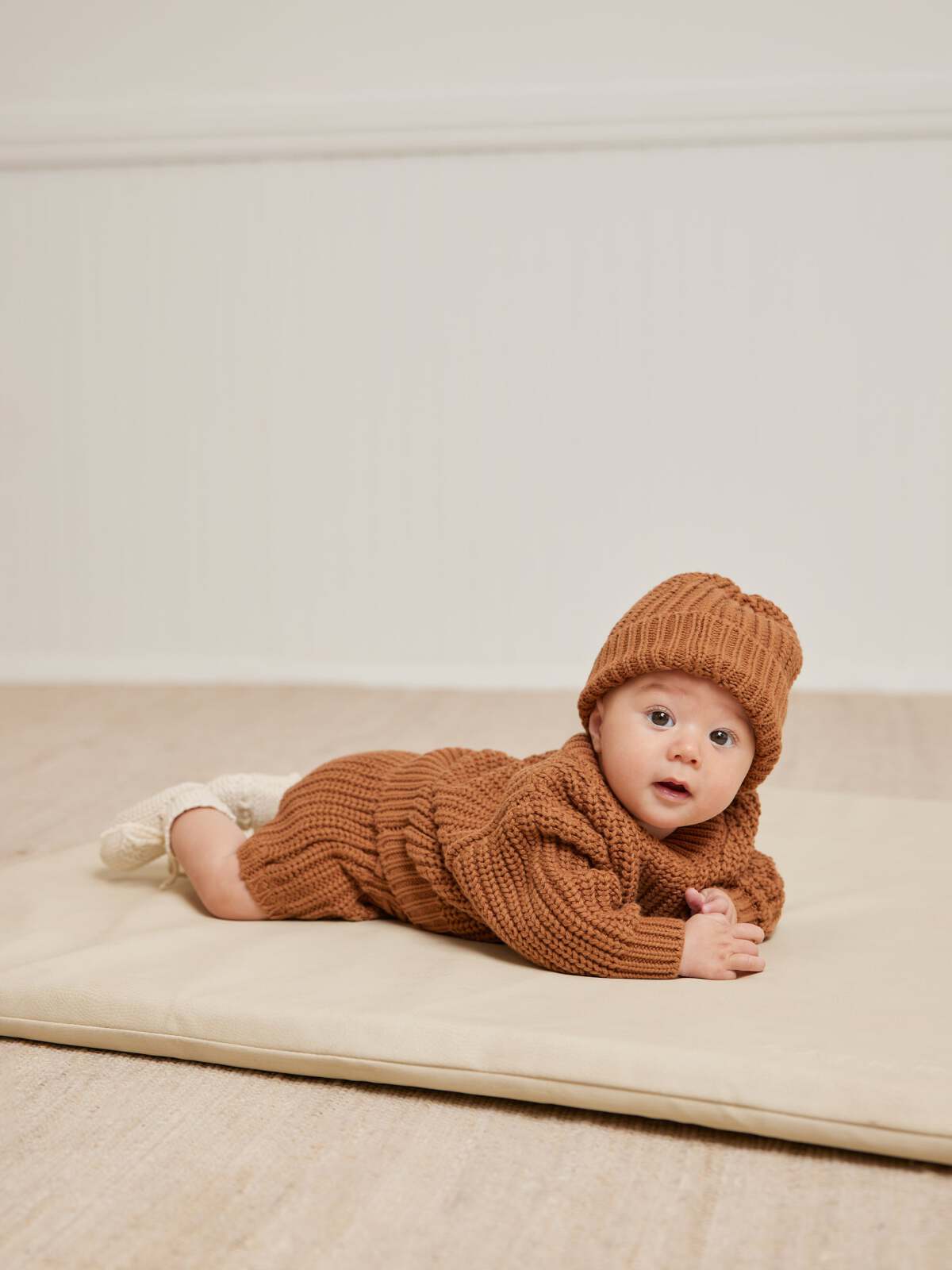 Baby in a brown knitted outfit and hat, lying on a soft mat, looking curiously at the camera.