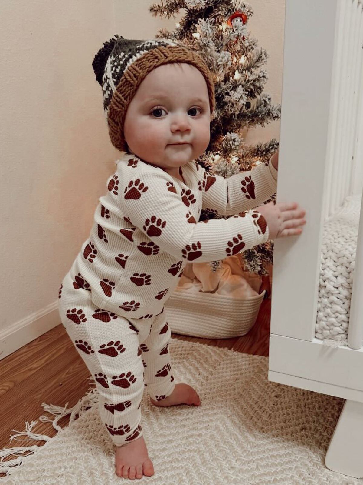 Baby in bear-print pajamas stands by a crib, smiling, next to a small decorated Christmas tree.