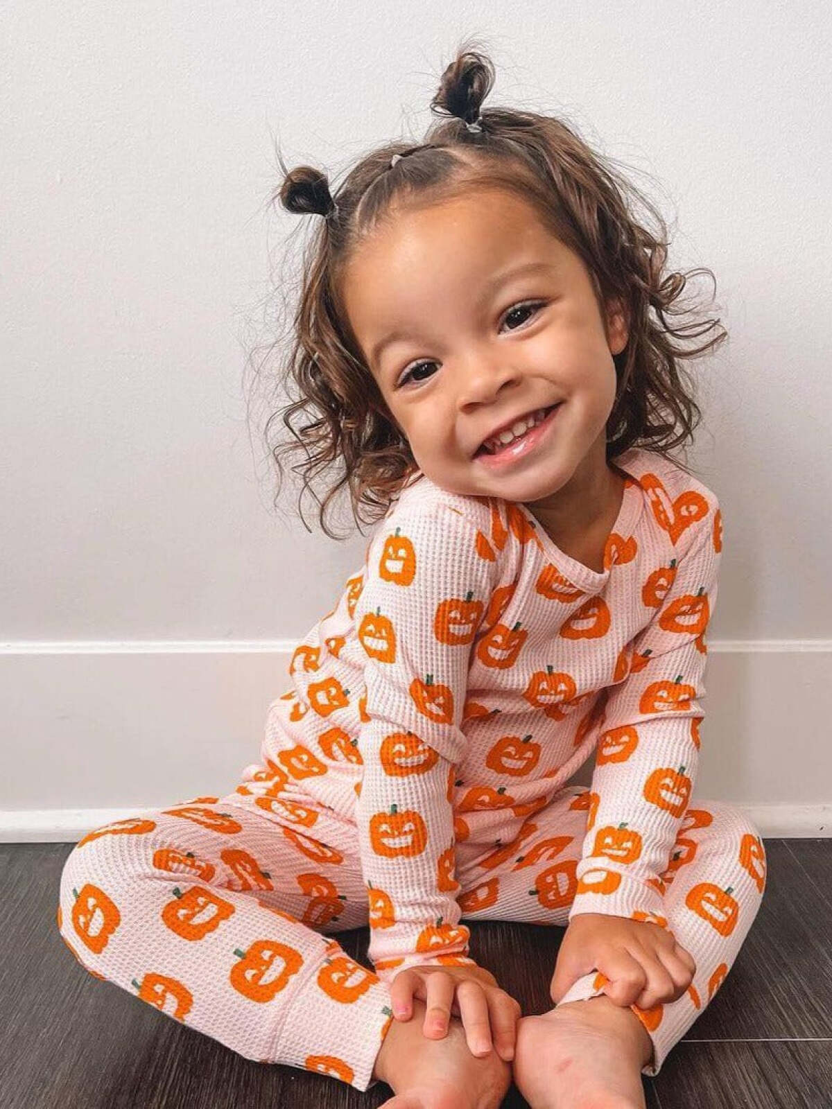 Smiling child in pumpkin-patterned pajamas sitting on the floor, with playful curly hair styled in two buns.