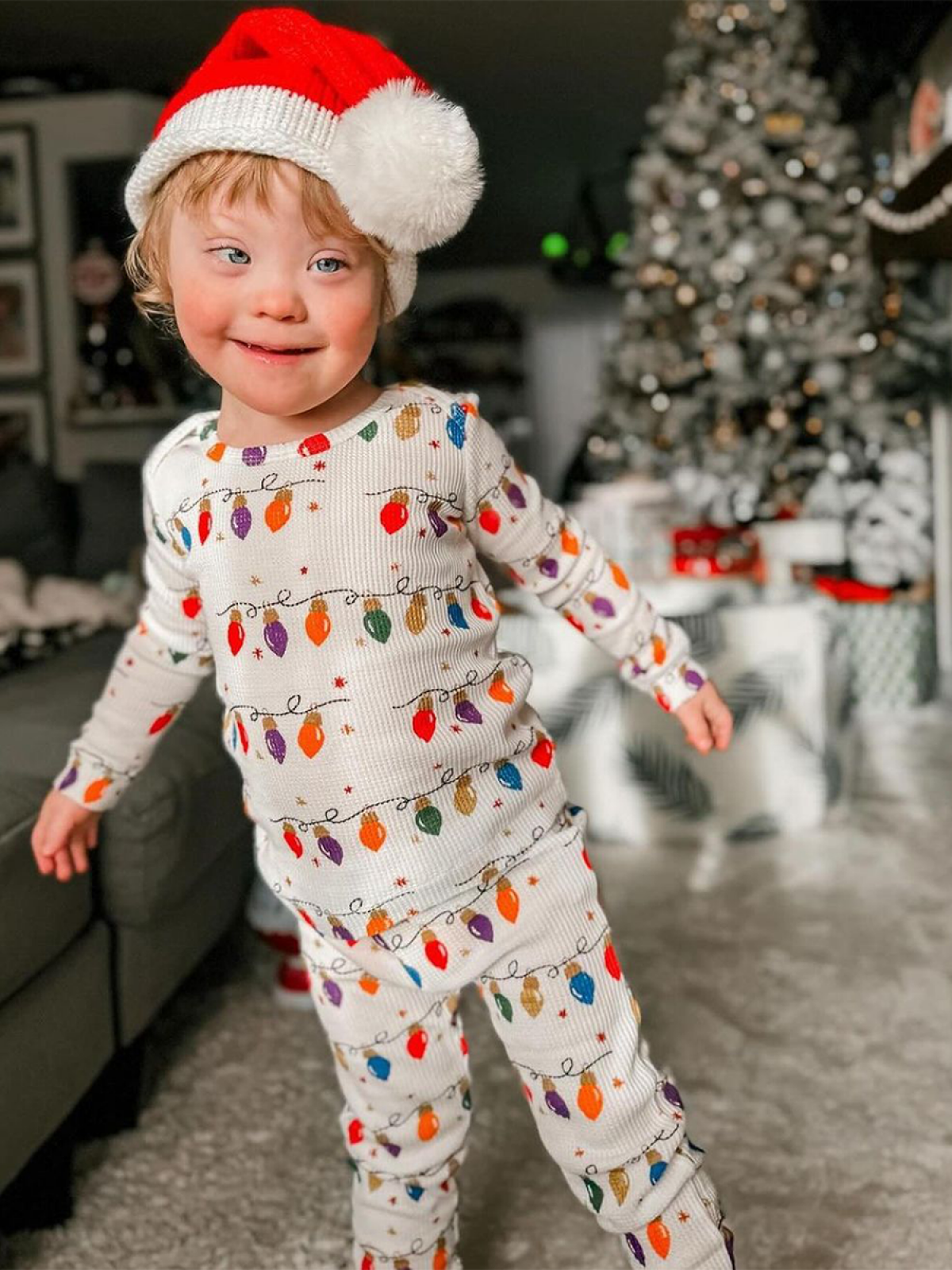 Child in festive pajamas with Christmas lights, wearing a Santa hat, smiling in a decorated living room.