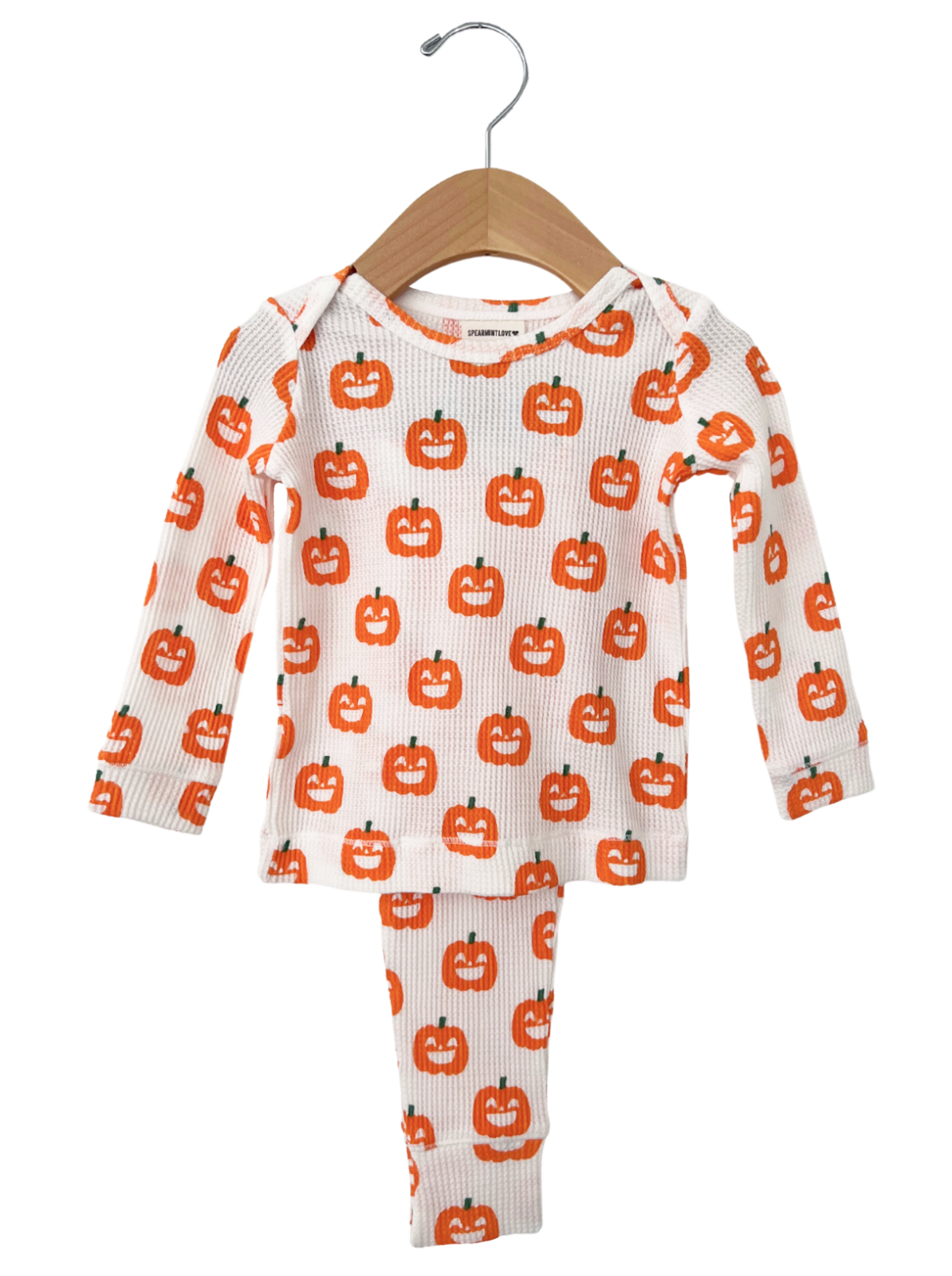 White pajama set with orange pumpkin faces, featuring a long-sleeve top and matching pants, hanging on a wooden hanger.