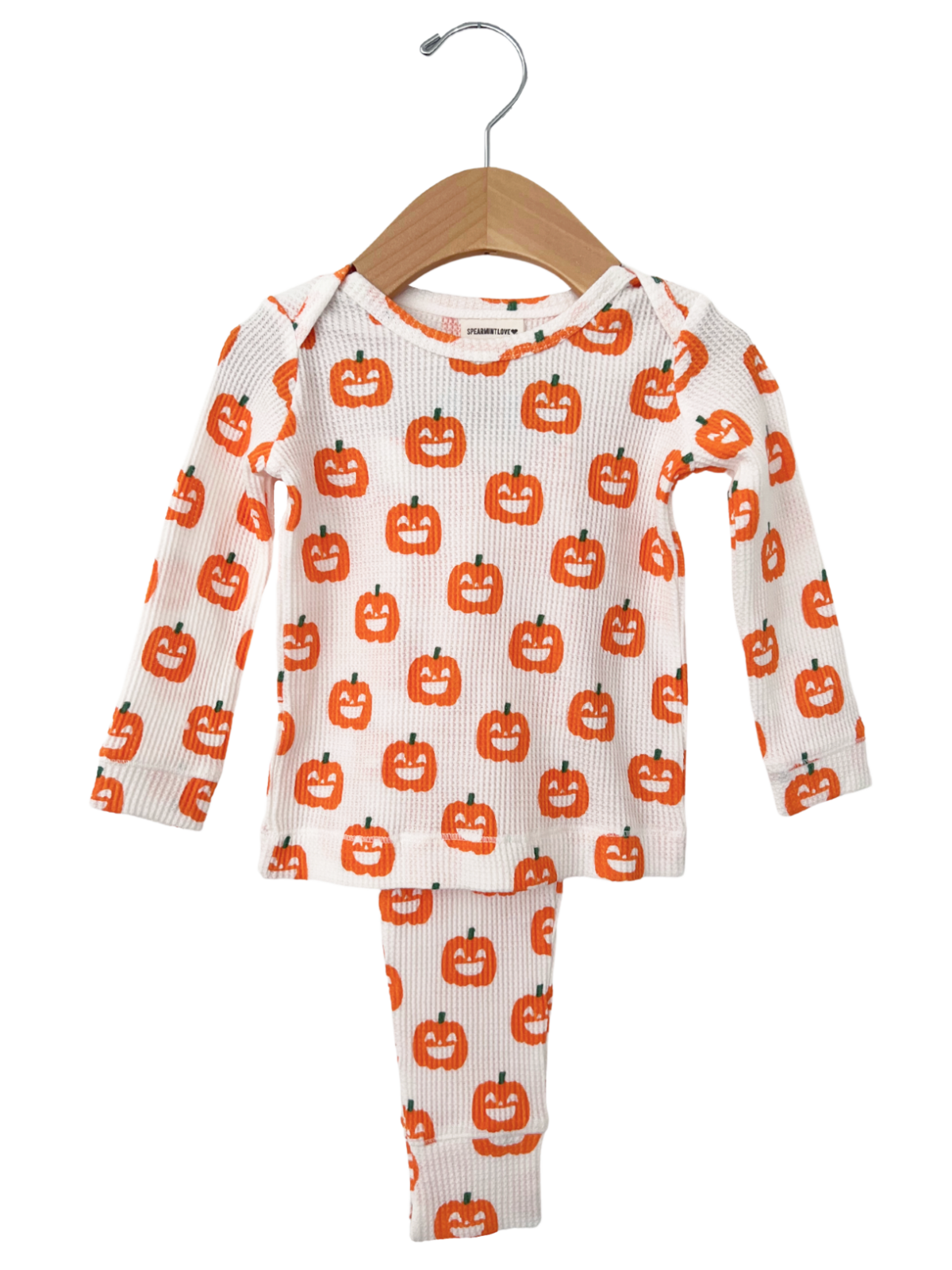 White pajama set with orange pumpkin faces, featuring a long-sleeve top and matching pants, hanging on a wooden hanger.