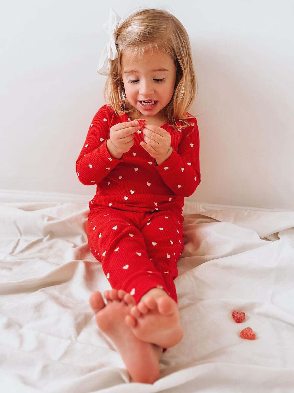 Child in red heart-patterned pajamas smiles while holding candy on a white blanket, with feet visible.
