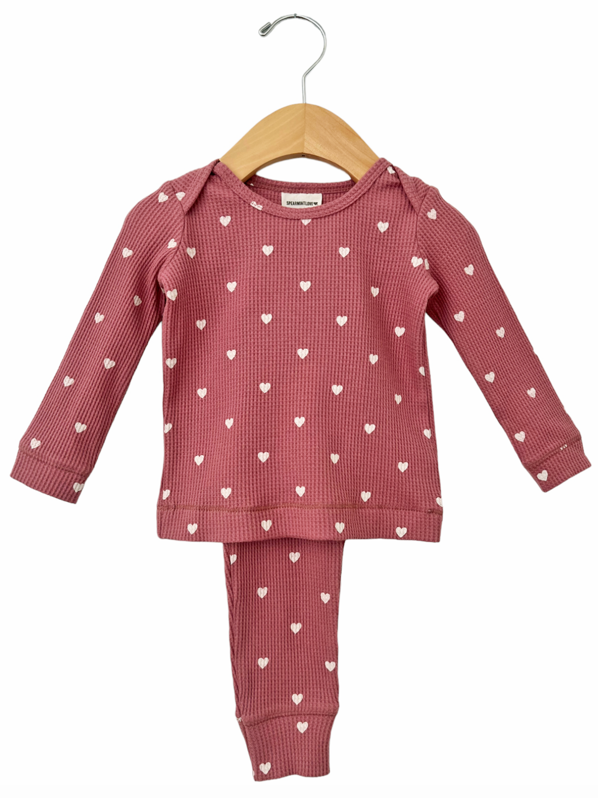 Pink toddler outfit with long sleeves and heart patterns, hanging on a wooden hanger against a white background.