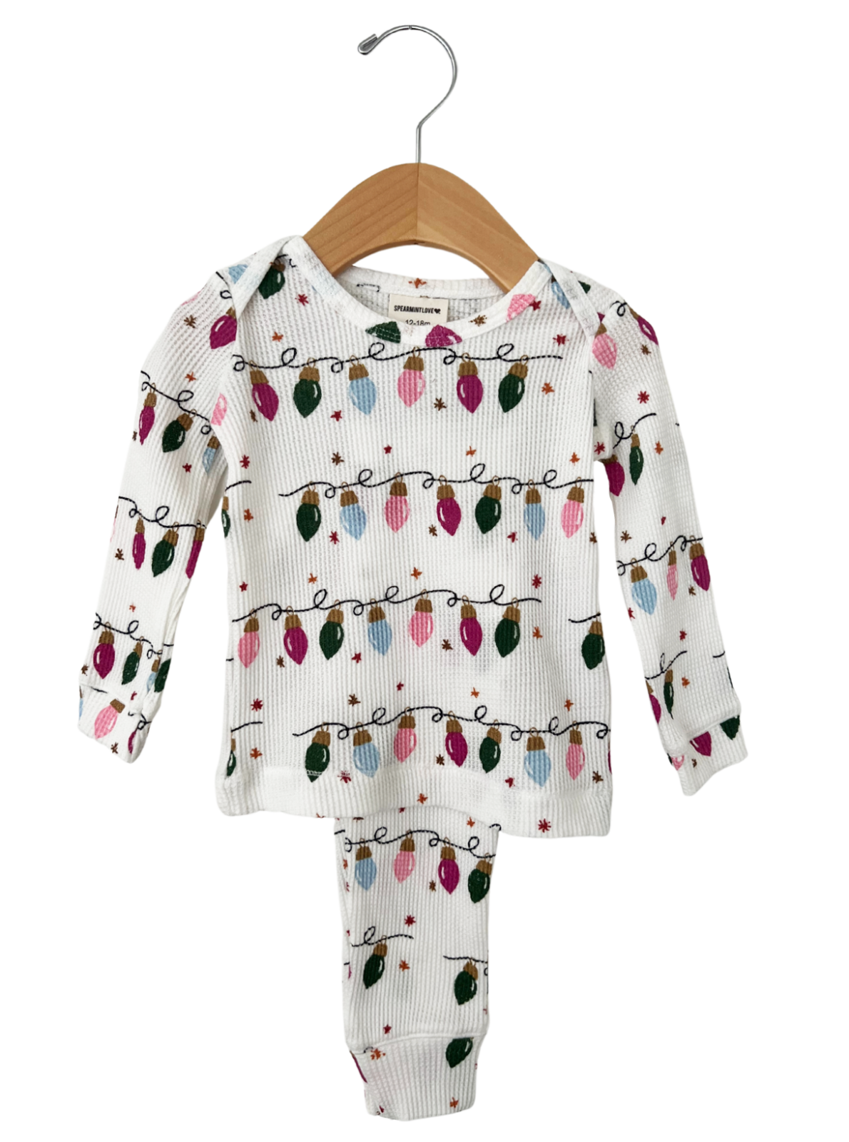 White pajamas with colorful Christmas lights pattern, hanging on a wooden hanger against a white background.