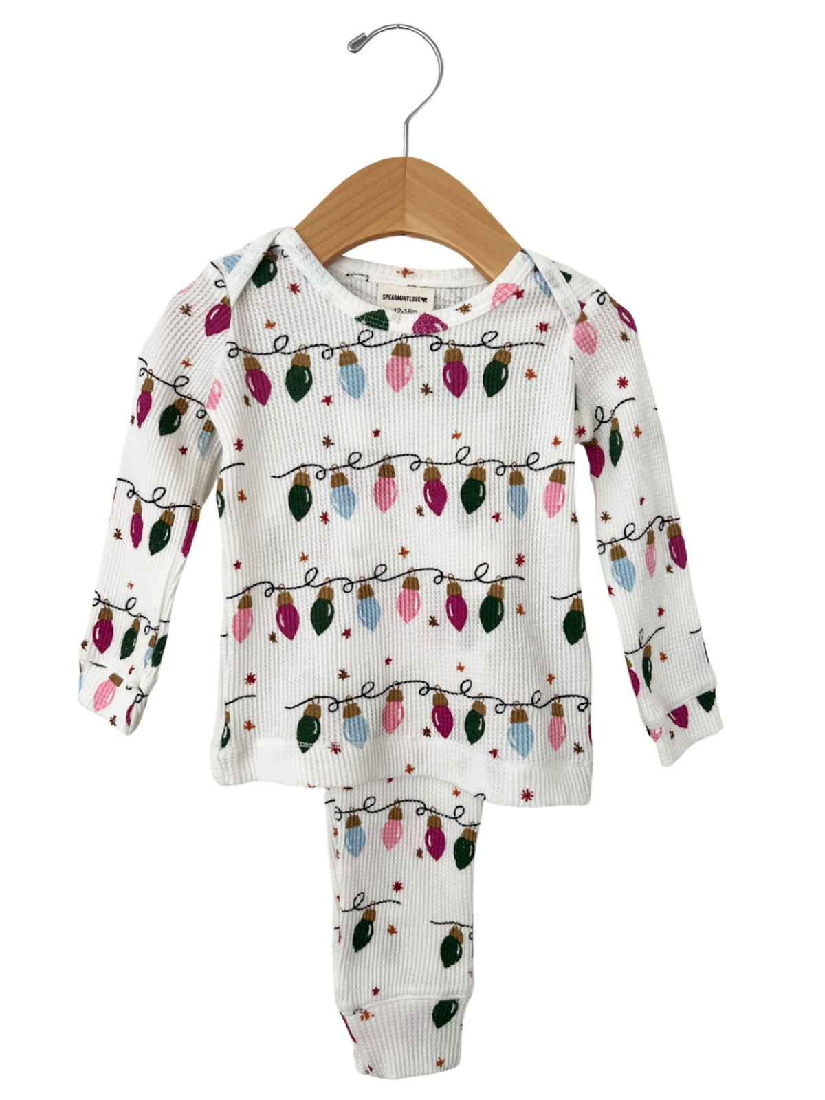 White pajamas with colorful Christmas lights pattern, hanging on a wooden hanger against a white background.