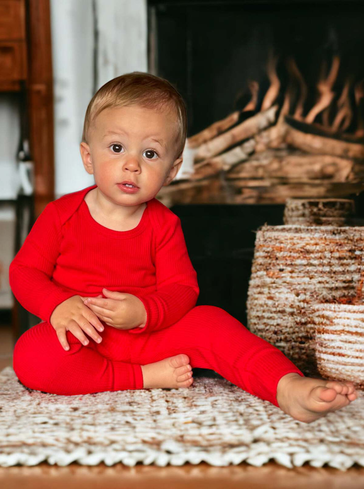 Toddler in red outfit sitting on woven rug near a fireplace, looking curiously at the camera.