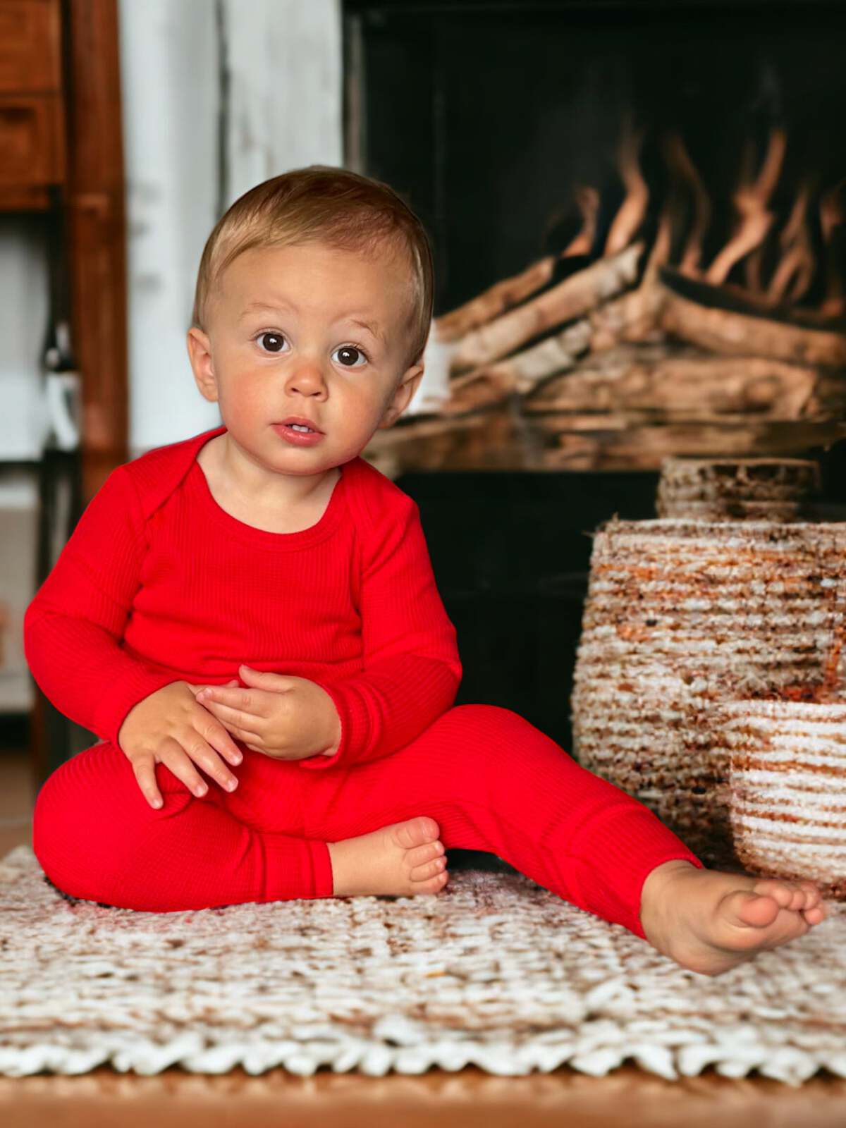 Toddler in red outfit sitting on woven rug near a fireplace, looking curiously at the camera.