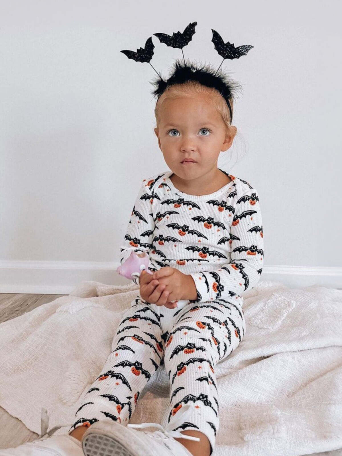 Toddler in bat-patterned outfit with bat headband, seated on a blanket, looking curiously at the camera.