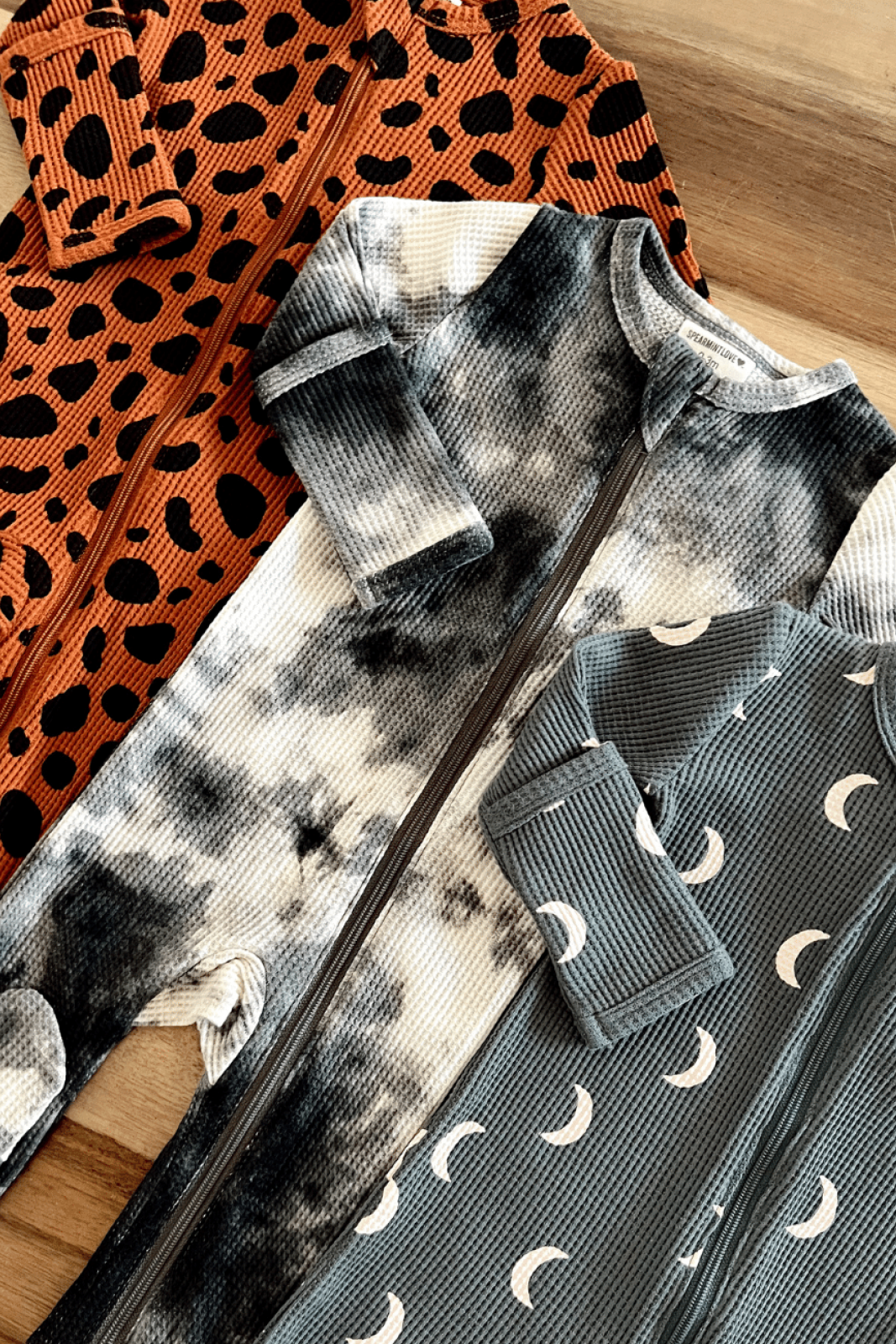 Three cozy baby onesies in animal print, tie-dye, and crescent moon designs, displayed on a wooden surface.