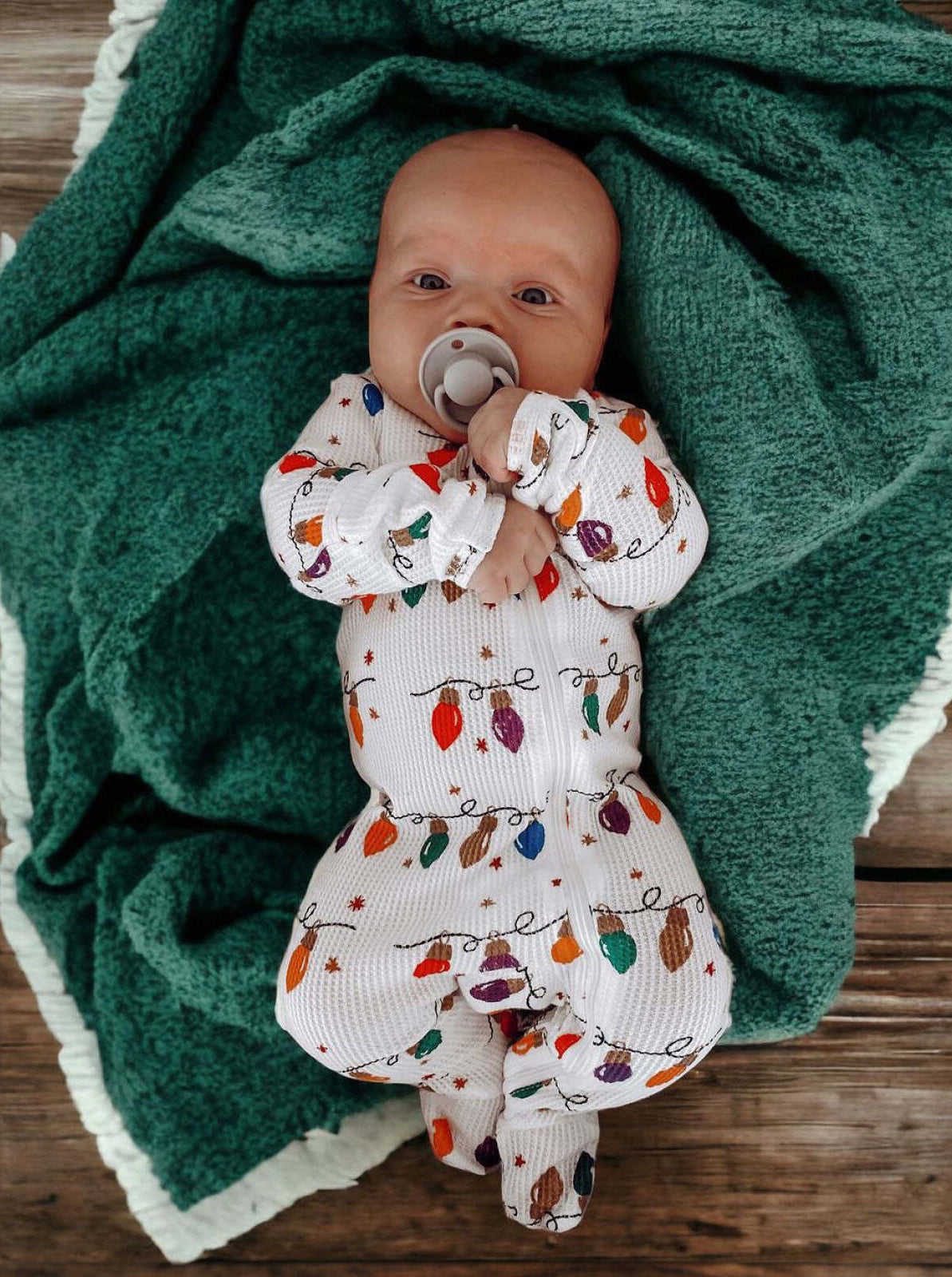 Baby in festive pajamas with colorful lights, lying on a green blanket, holding a pacifier.