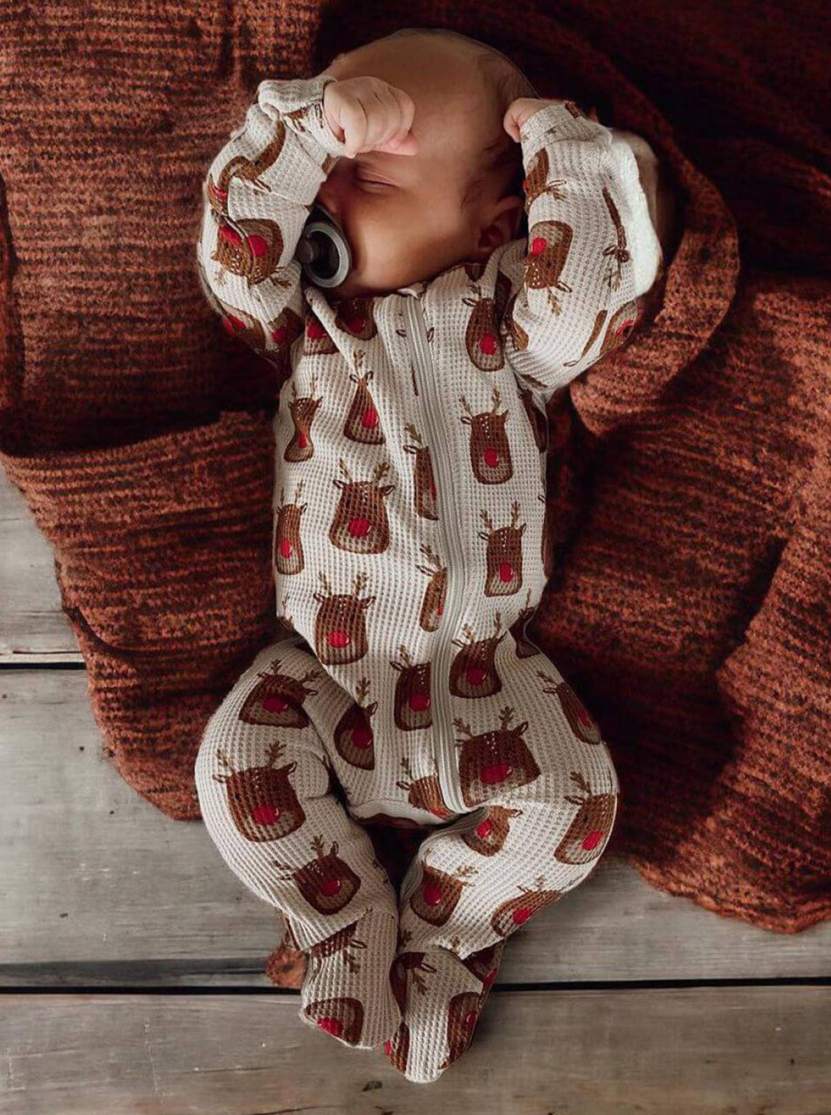 Baby in a reindeer-patterned onesie resting on a cozy, textured rust blanket.