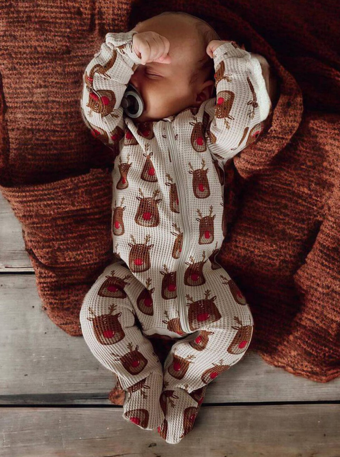 Baby in a reindeer-patterned onesie resting on a cozy, textured rust blanket.