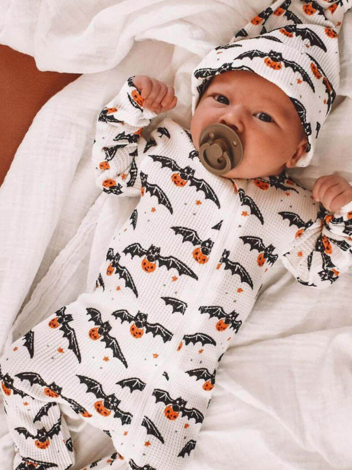 Baby in a cozy Halloween-themed outfit with bats and pumpkins, lying on a blanket.