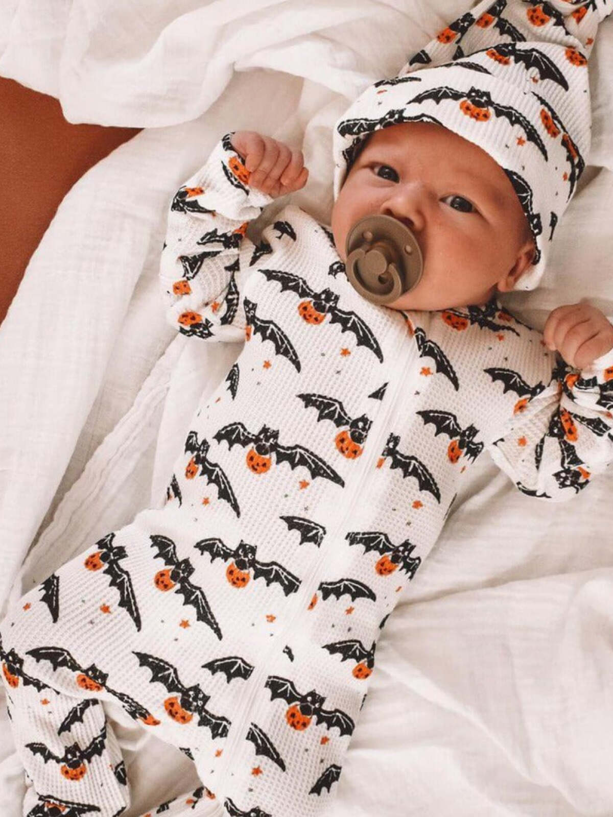 Baby in a cozy Halloween-themed outfit with bats and pumpkins, lying on a blanket.