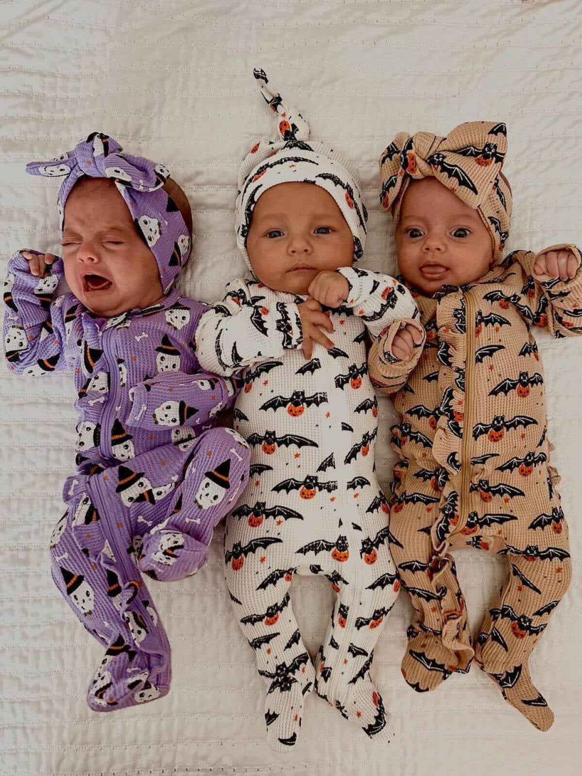 Three babies in Halloween-themed outfits: one crying in purple, one smiling in white with bats, and one smiling in tan.