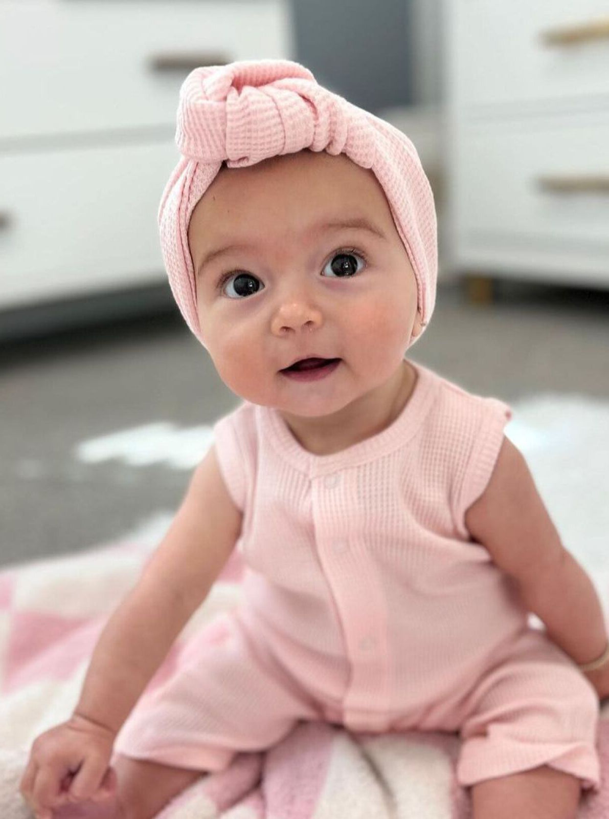 Smiling baby girl in a pink outfit and headband, seated on a blanket in a bright room.