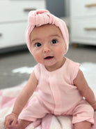 Smiling baby girl in a pink outfit and headband, seated on a blanket in a bright room.