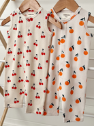 Two baby rompers hang on wooden hangers, featuring cherry and orange patterns on a light background.