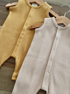 Two sleeveless, ribbed onesies in mustard and beige hanging on wooden hangers against a wooden surface.