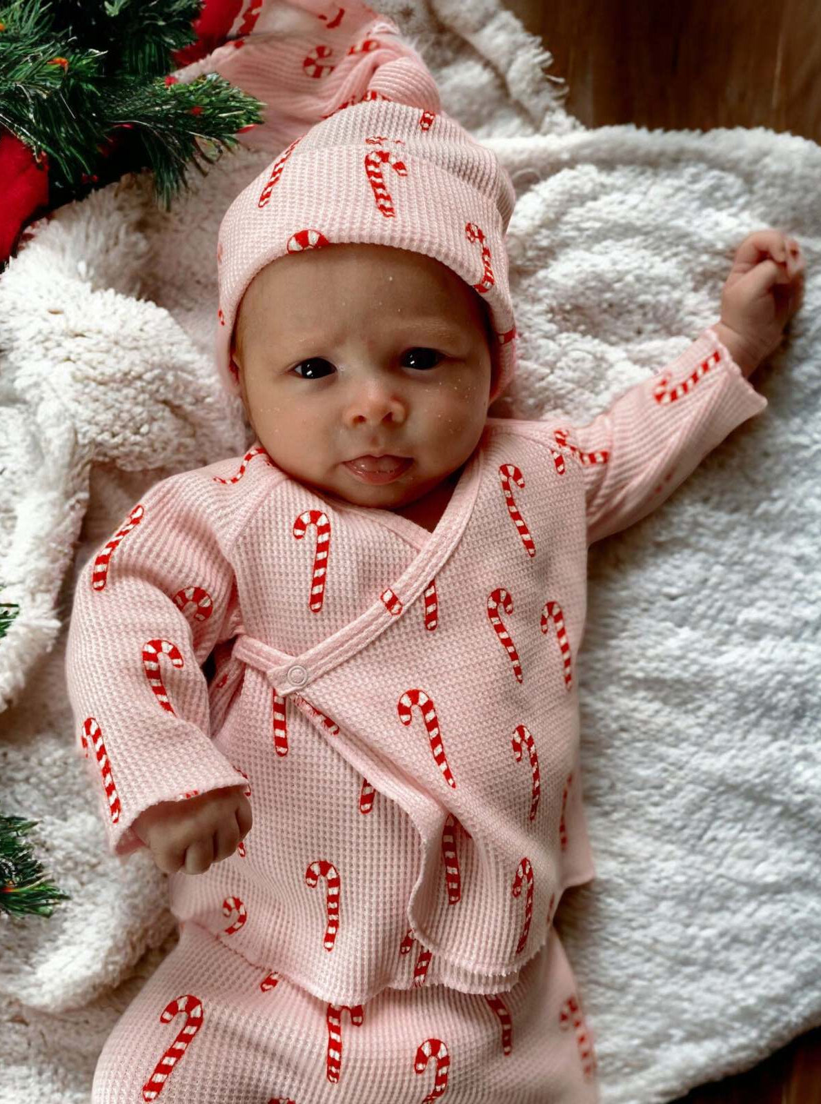 Baby in candy cane pajamas lying on a white blanket, surrounded by festive greenery.