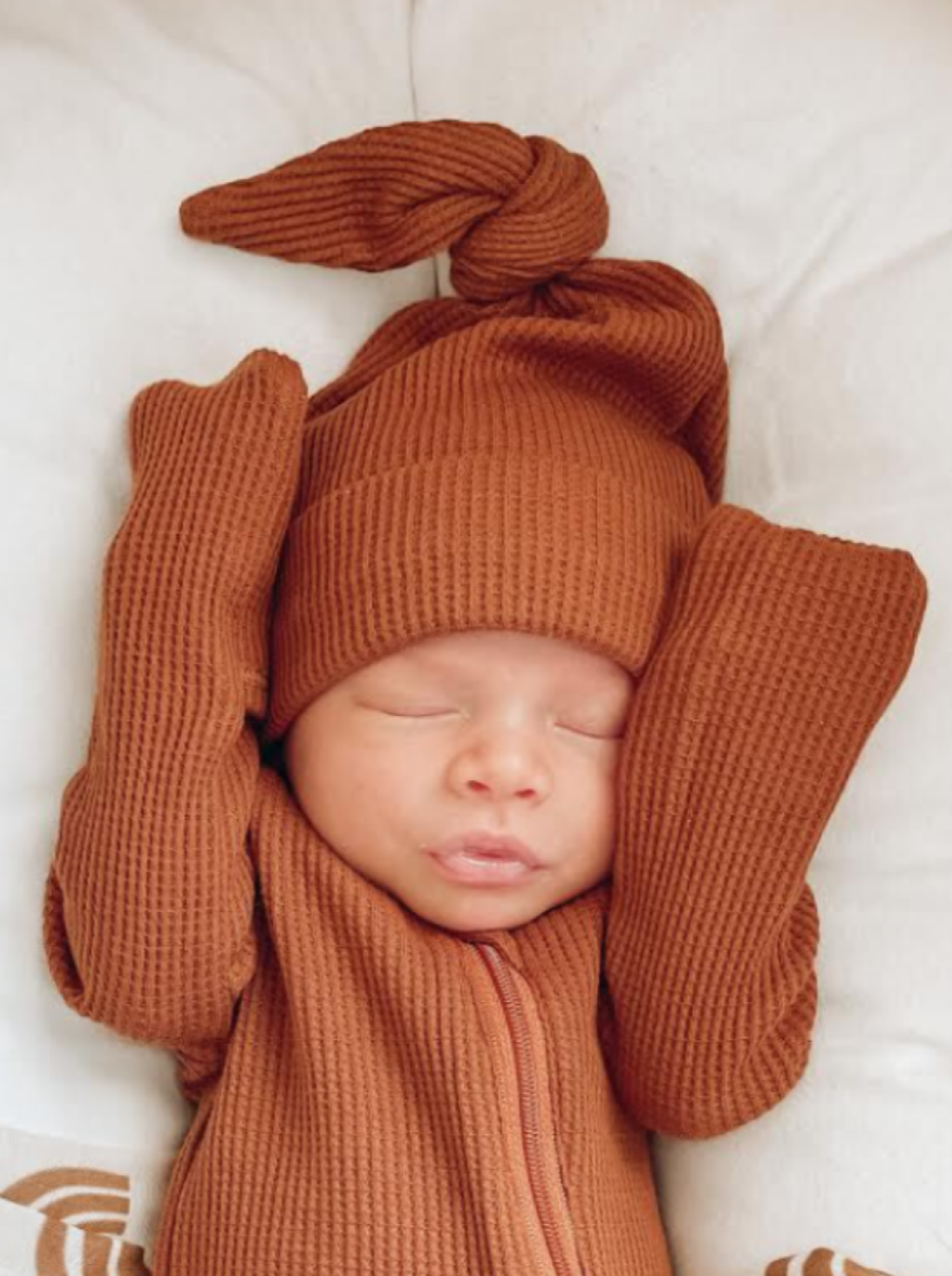 Sleeping baby in a brown ribbed outfit with a knotted hat, resting peacefully on a soft surface.