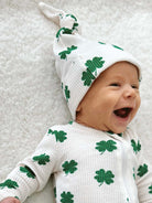 Smiling baby in a white outfit with green clovers and a matching hat against a soft background.