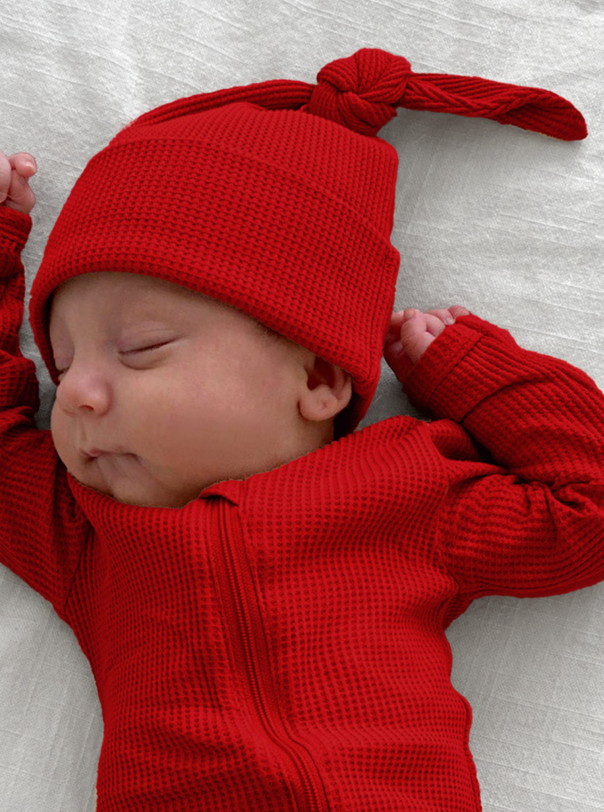Sleeping baby in a red outfit and hat, resting on a white blanket.