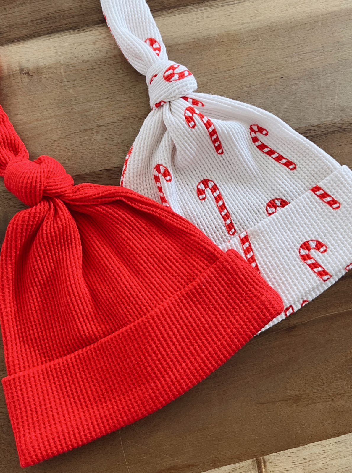 Red and white patterned baby hats with candy cane design, tied at the top, on a wooden surface.
