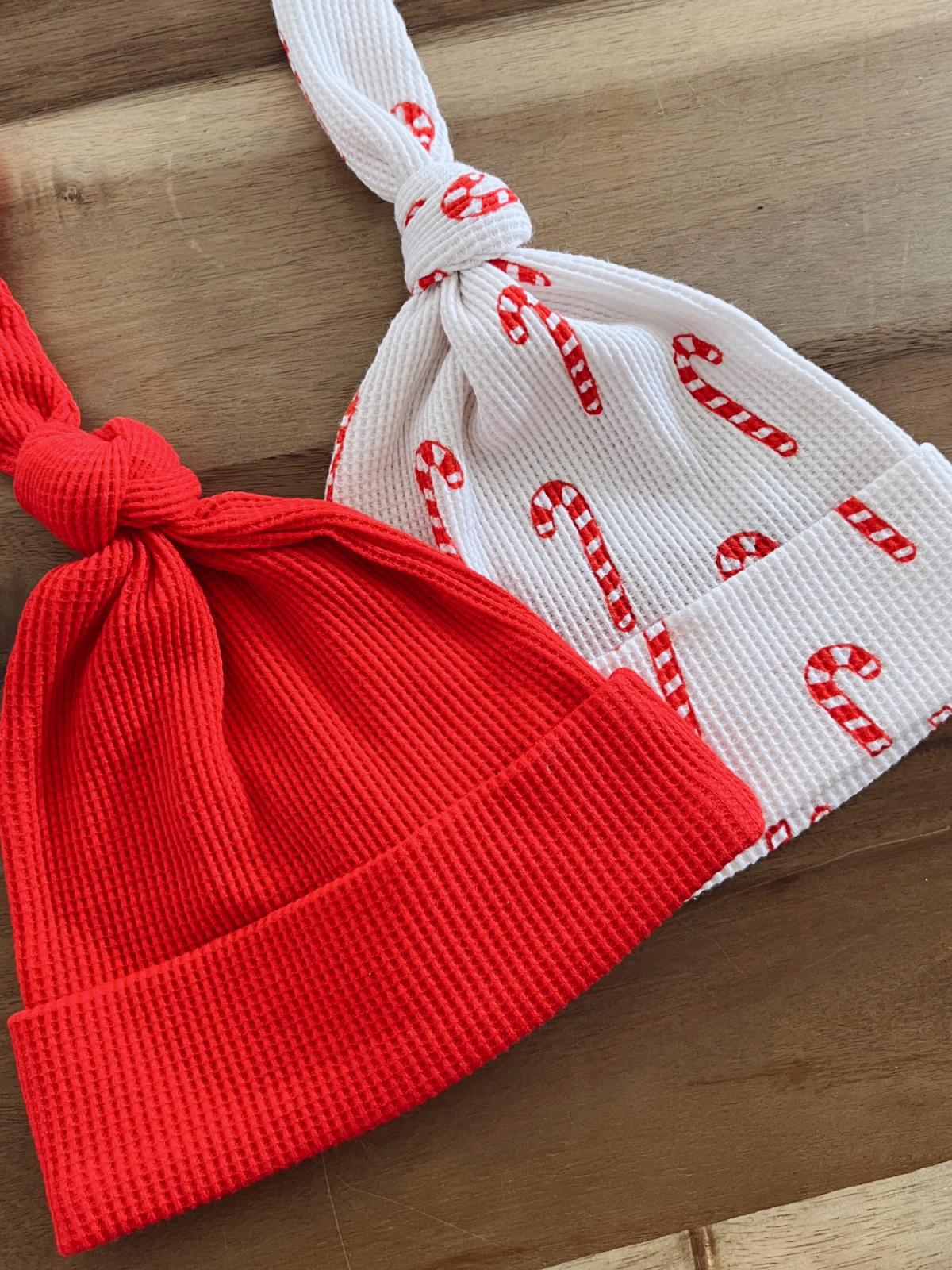 Red and white patterned baby hats with candy cane design, tied at the top, on a wooden surface.