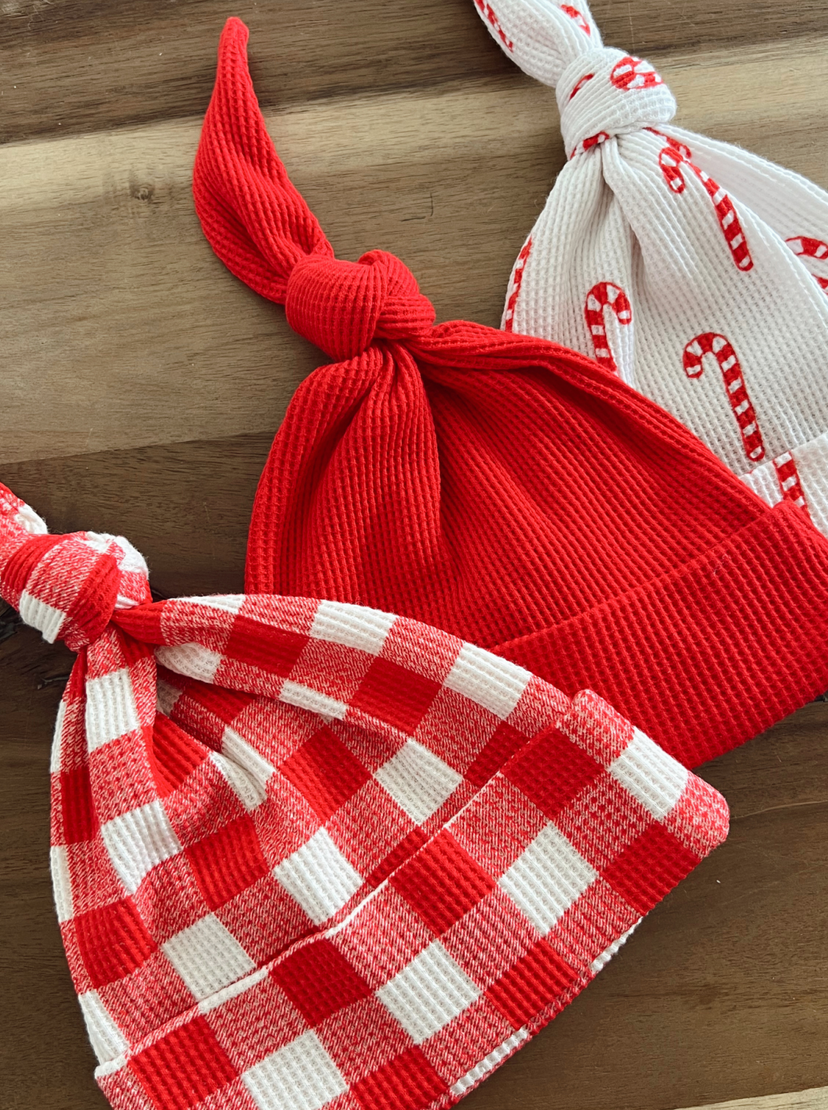 Three festive hats: a solid red, a red and white checkered, and a white with candy cane pattern on a wooden surface.