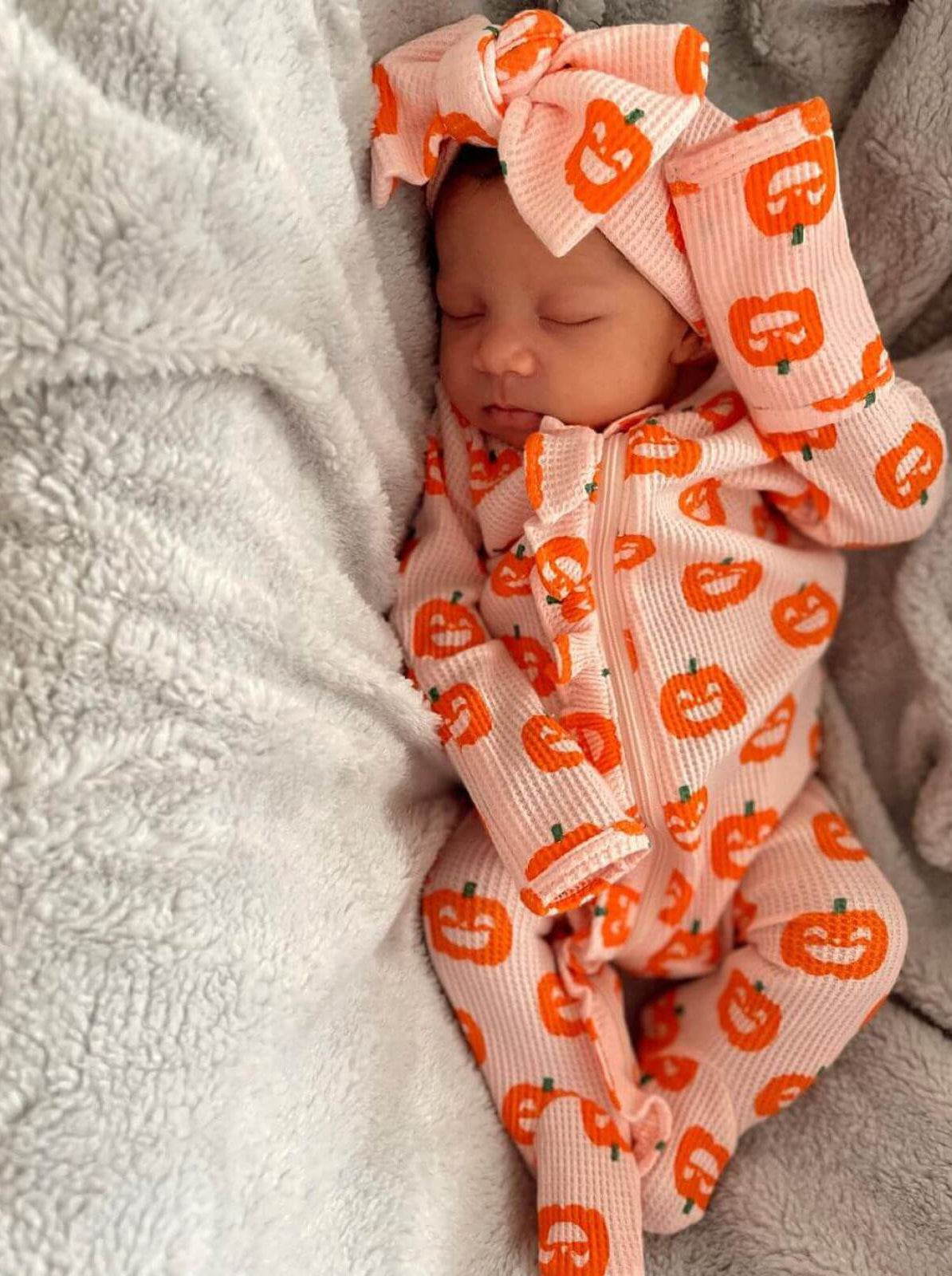 Sleeping baby in pumpkin-patterned pajamas, nestled in soft gray blankets.