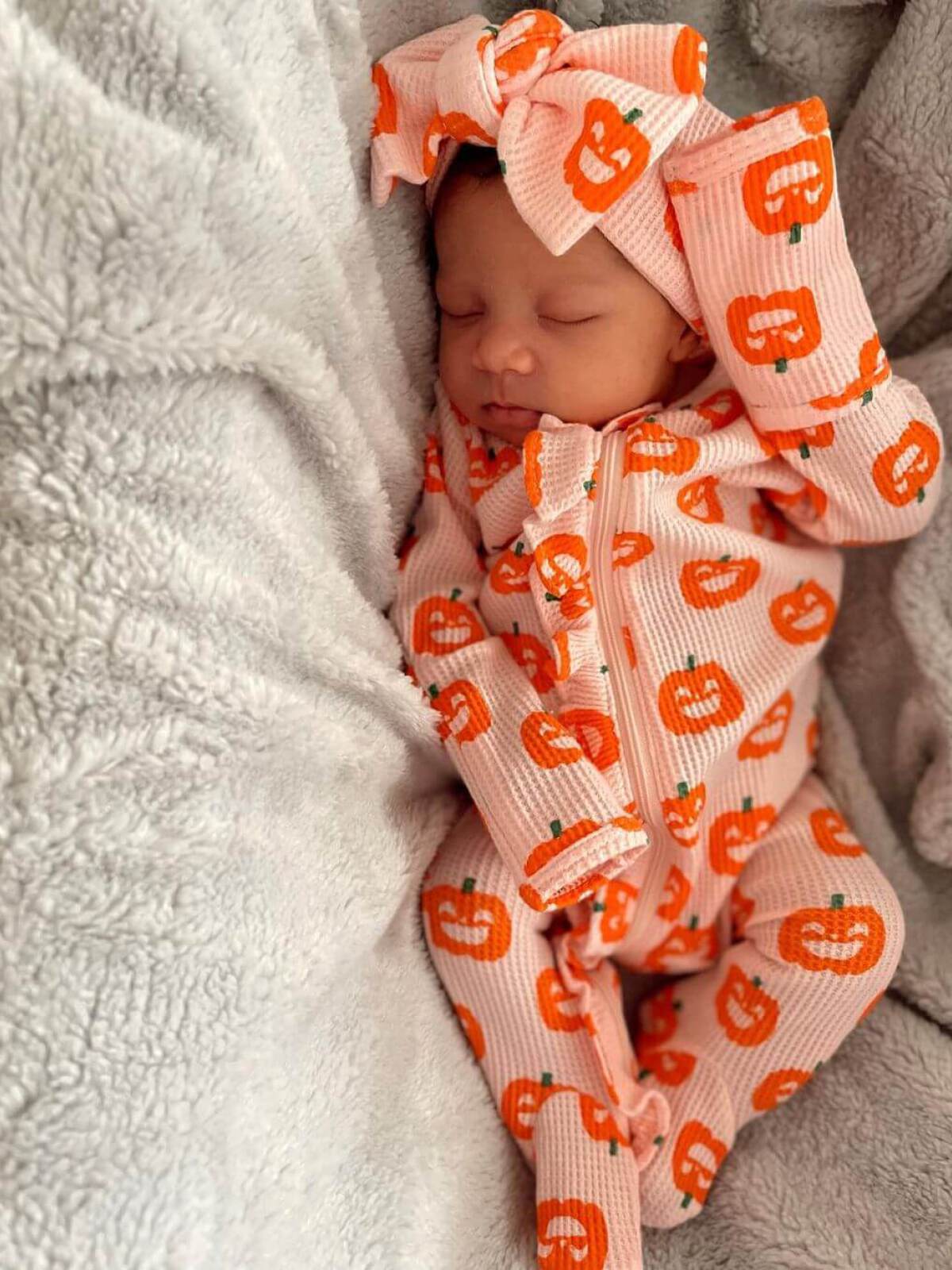 Sleeping baby in pumpkin-patterned pajamas, nestled in soft gray blankets.