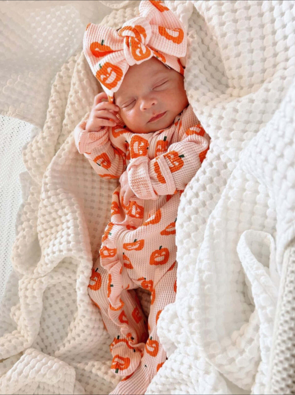 Sleeping baby in pumpkin-patterned outfit with a large bow, nestled in a cozy white blanket.