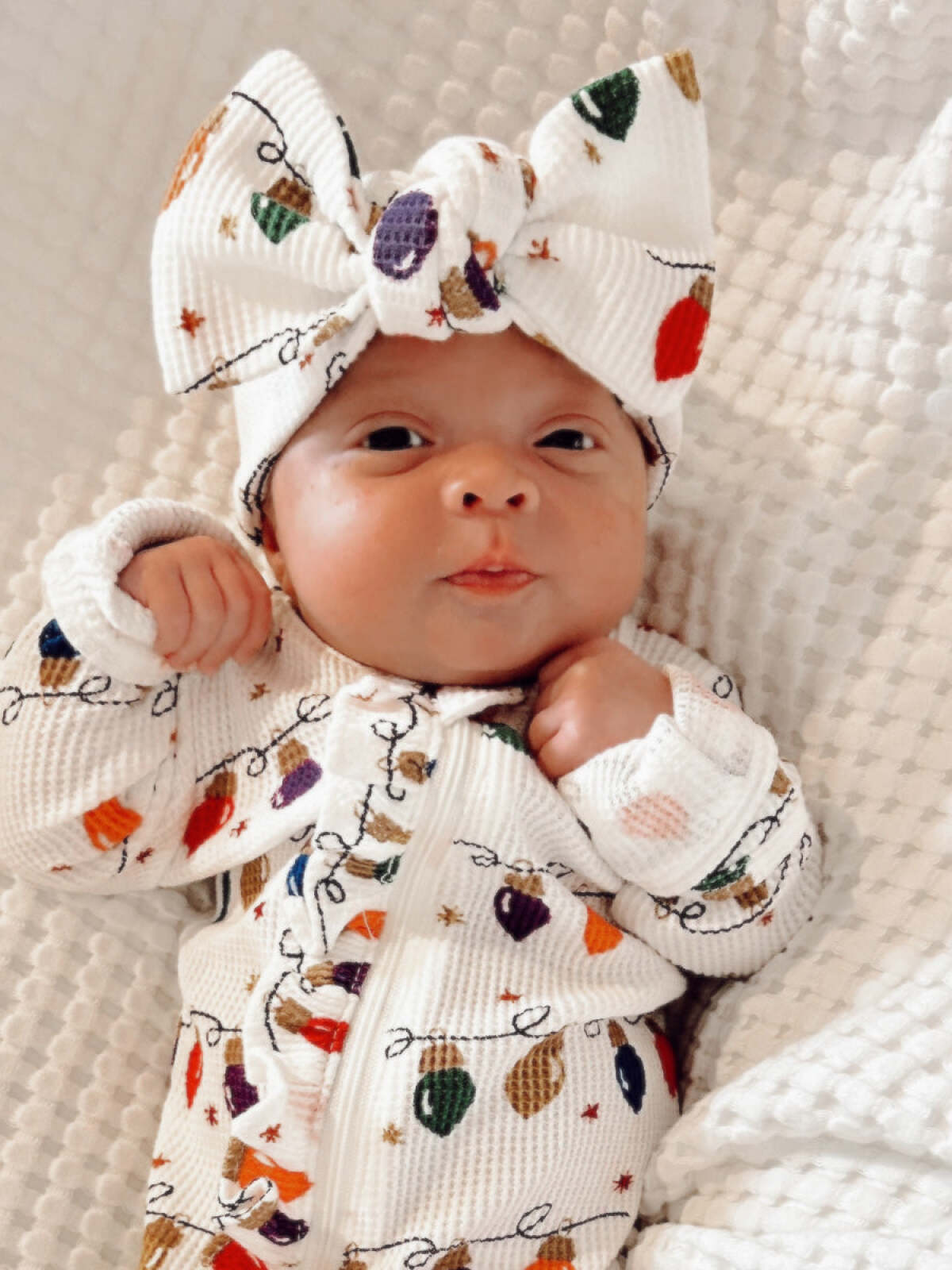 Baby in festive outfit with holiday lights design and a large bow, lying on a textured white blanket.