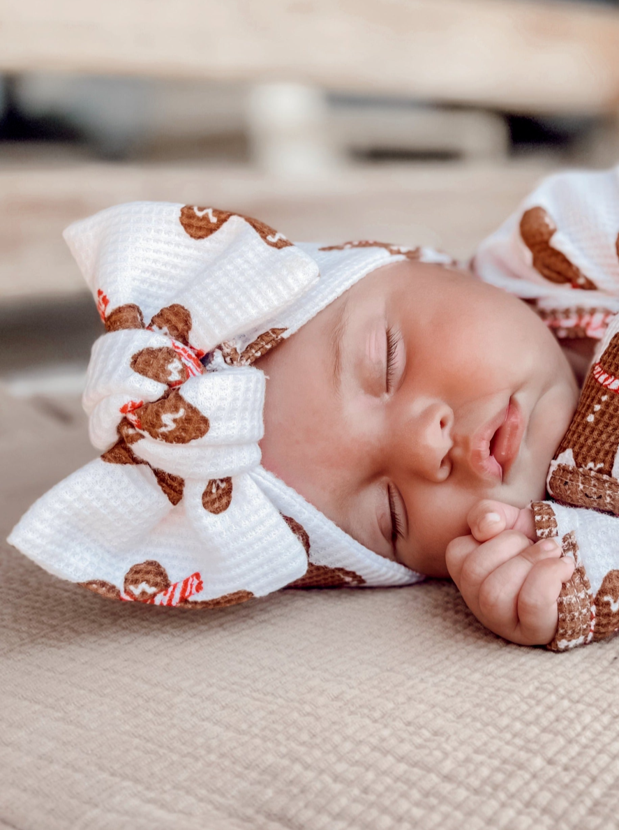Sleeping baby wearing a white and brown gingerbread-themed headband on a textured blanket.