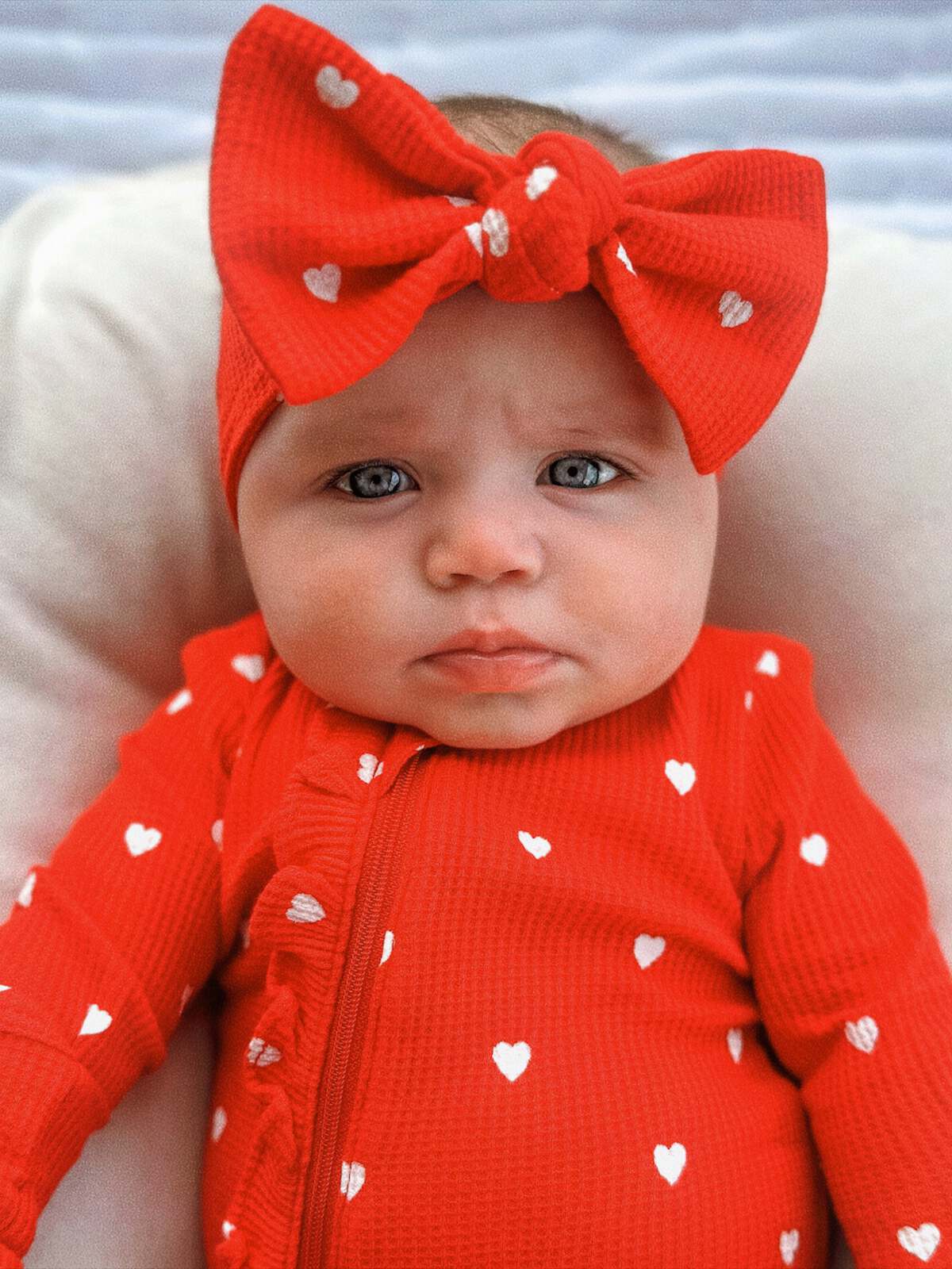 Baby with blue eyes wearing a red outfit and large bow, resting on a soft surface.