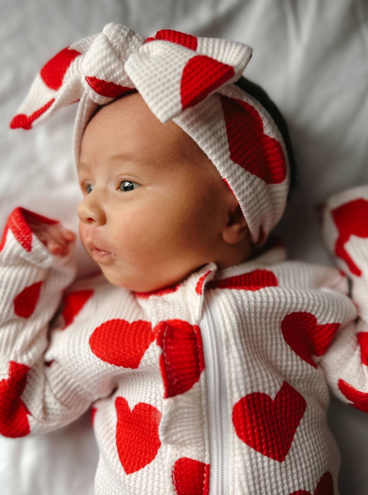 Infant in a heart-patterned outfit and headband, lying on a white blanket, looking thoughtfully to the side.