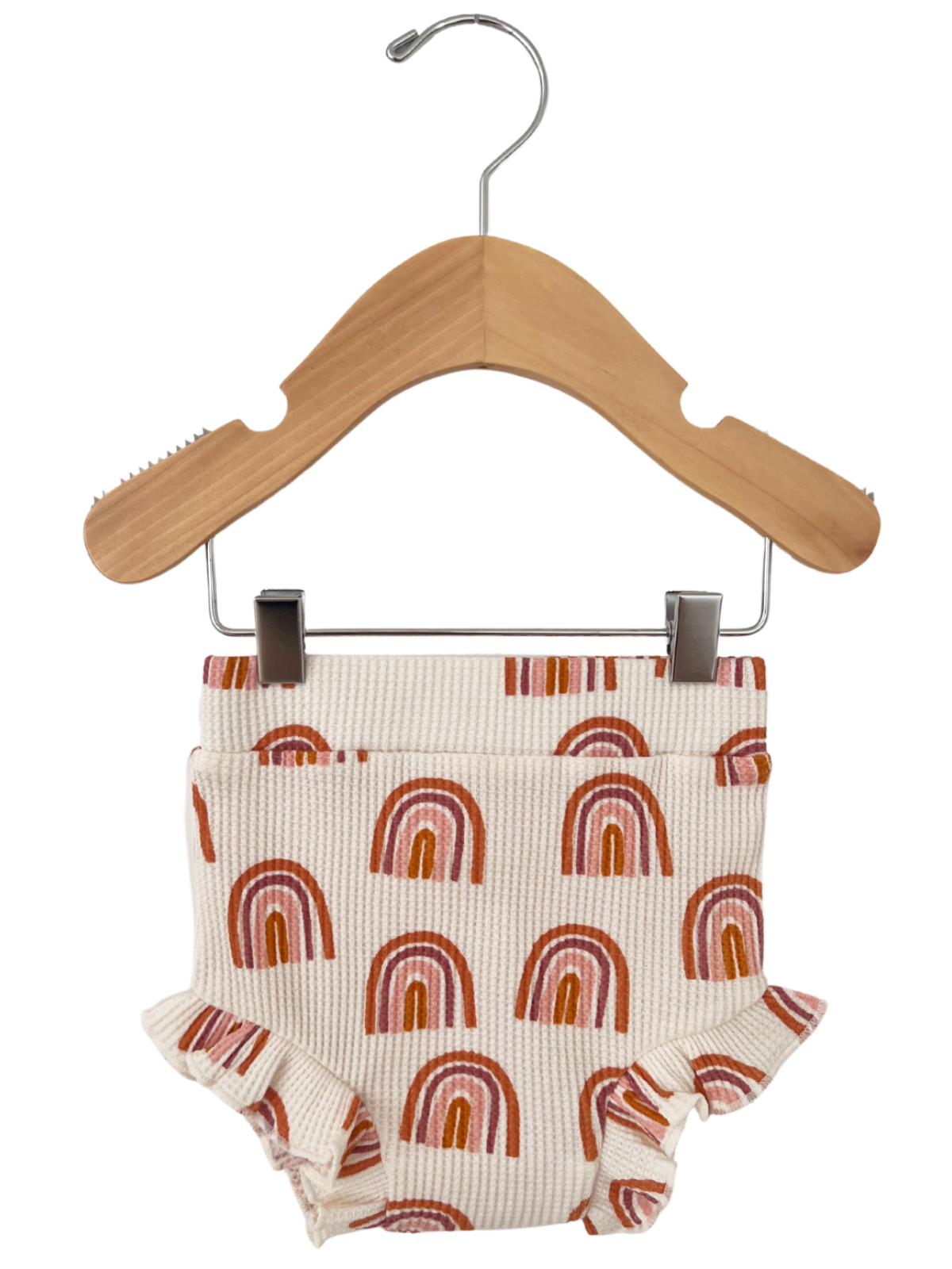 Baby shorts with rainbow pattern, ruffled edges, hanging on a wooden clothes hanger.