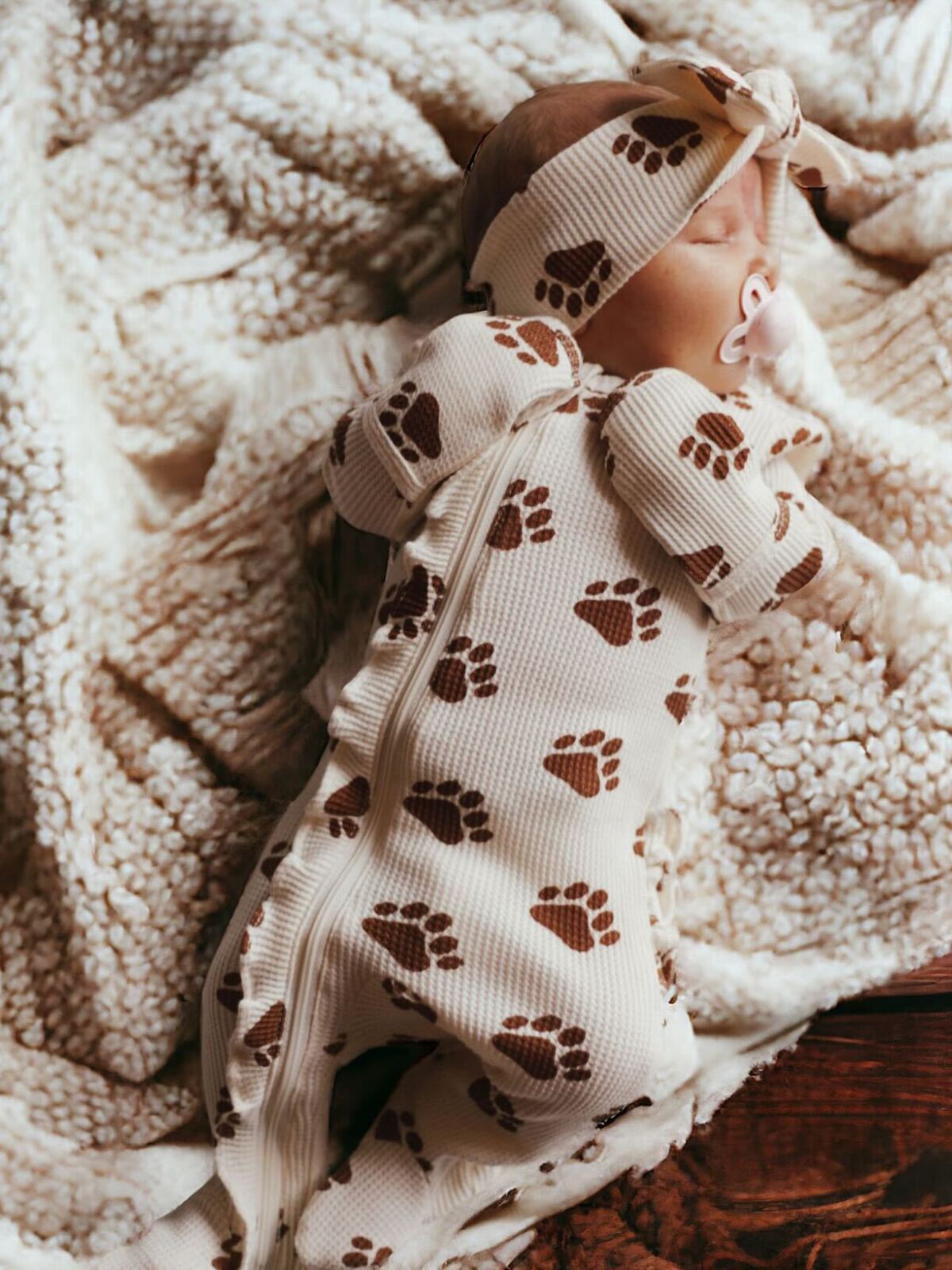 Newborn baby asleep on a cozy blanket, wearing a paw-print patterned onesie and matching headband.