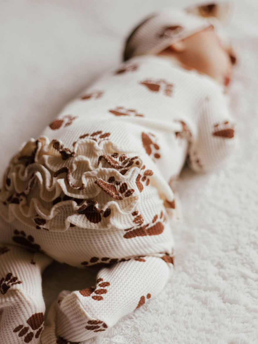 Baby peacefully sleeping on a soft blanket, wearing a white outfit with brown paw prints and a matching hat.