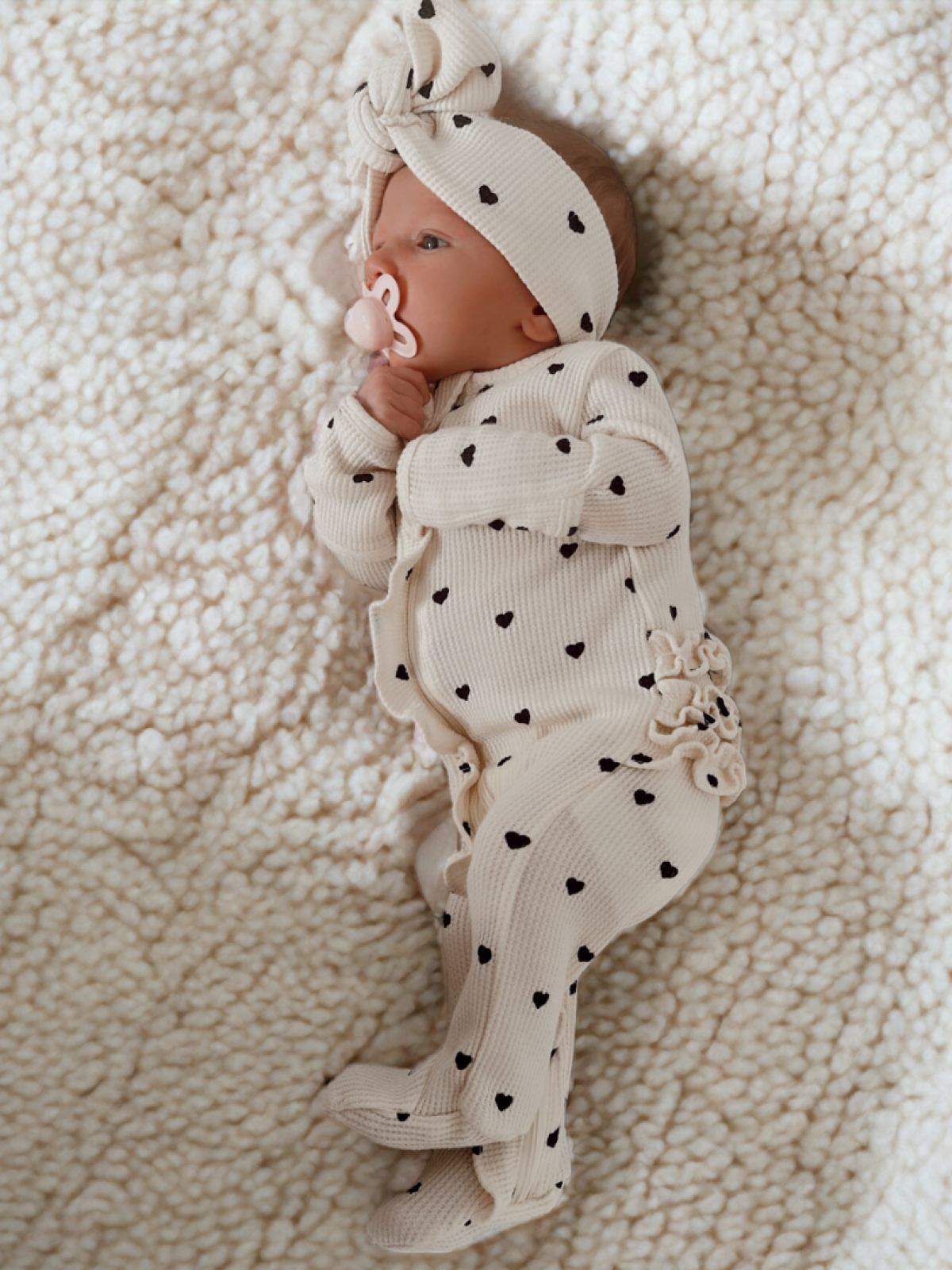 Infant in a heart-patterned onesie and headband, lying on a soft, textured blanket.